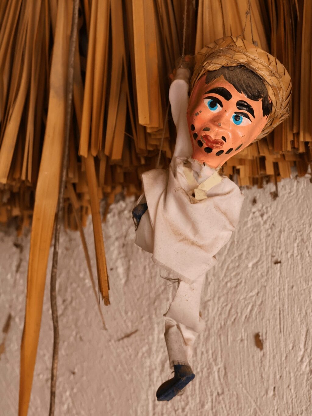 Eine kleine handgemachte Puppe mit weissen Stoffkleidern, schwarzen Schuhen und einem Strohhut hängt an einer Strohhalm-Decke.